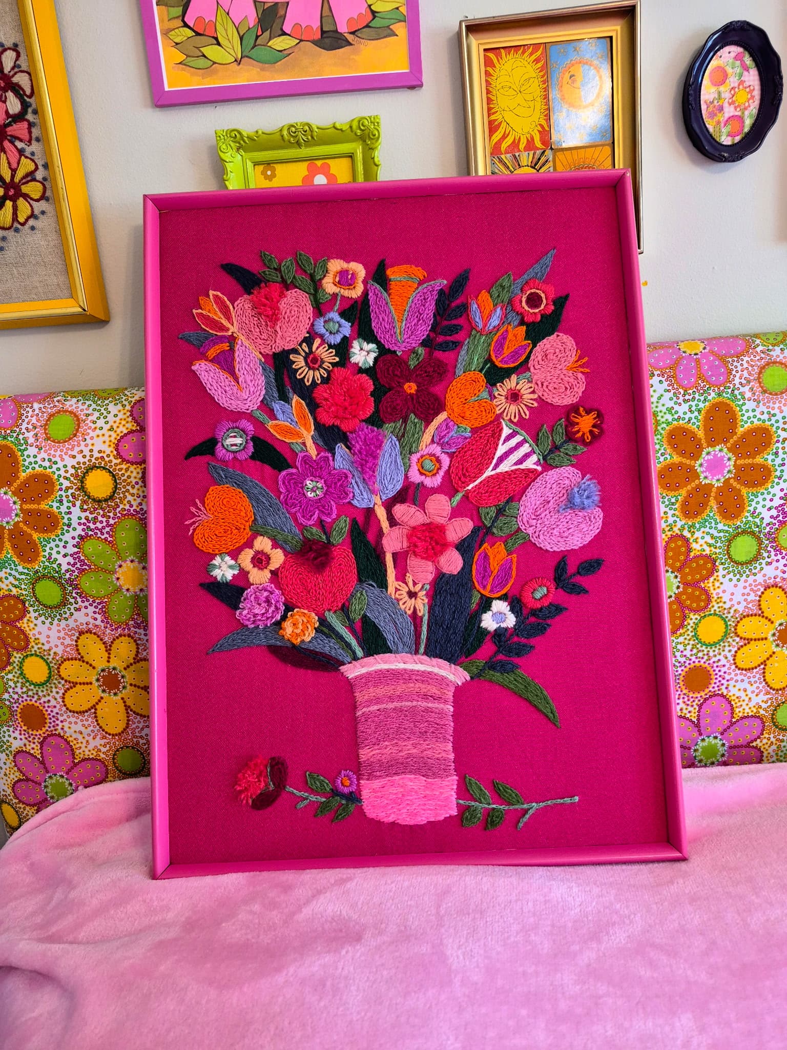 Restored vintage crewel embroidery floral bouquet in a hot magenta frame, styled against a vibrant retro backdrop with colorful floral pillows