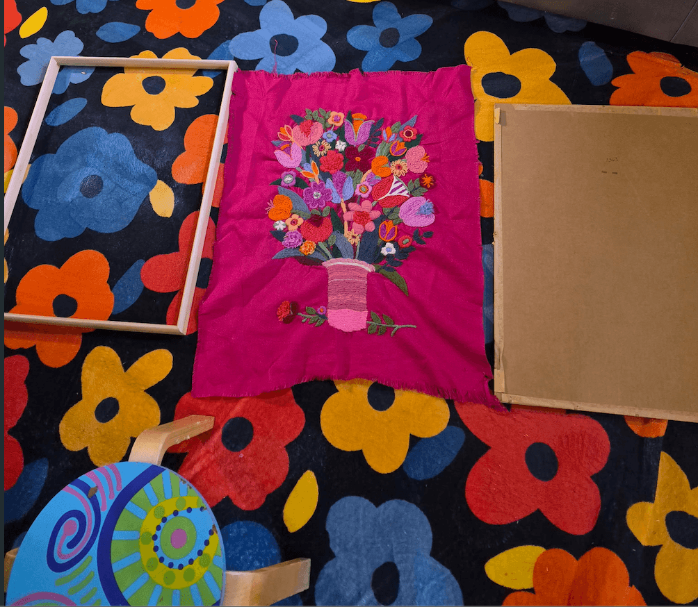 The same embroidery piece during restoration — removed from its original frame, laid on a bold retro floral fabric with the new pink frame being fitted